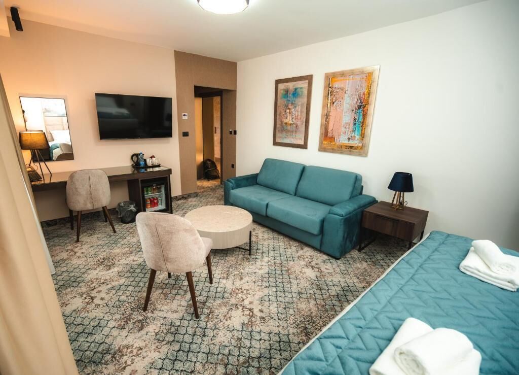 A hotel room with a sofa, a chair, a bed, a TV, artwork on the wall, and a small table with a lamp.