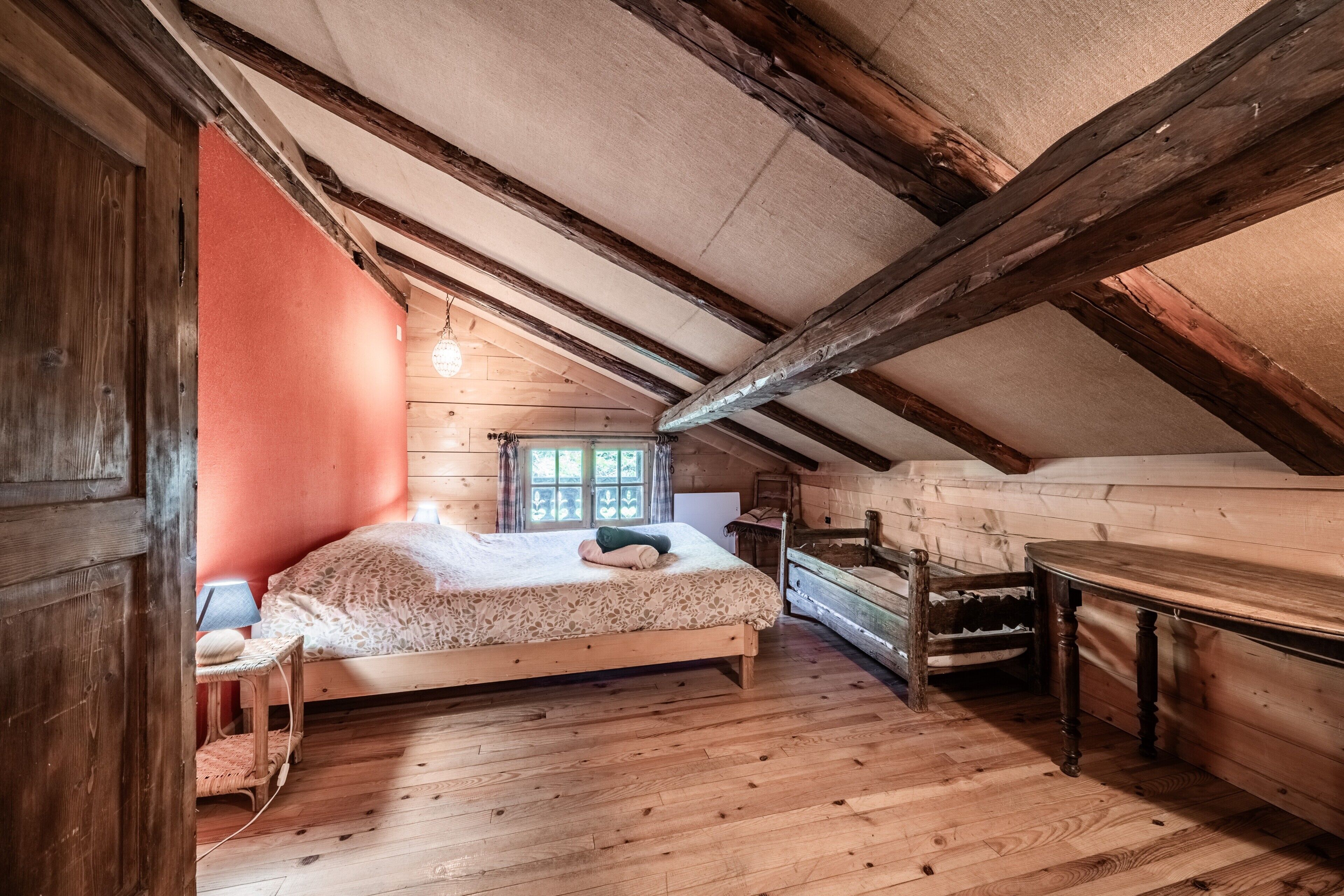 Ein Zimmer mit Holzwänden und schrägen Decken, einem Bett mit gemustertem Überwurf, einem kleinen Tisch und einem Fenster mit Vorhängen.
