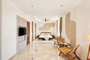Ein modernes Hotelzimmer mit Flachbildfernseher, einem Bett mit Holz Kopfteil, Deckenventilator und einer Sitzecke mit Stühlen und einem kleinen Tisch.
