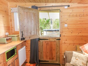 A compact kitchen with wooden walls, a window with a view, a microwave, a toaster, and a small refrigerator. - The Cabin (Amlwch)