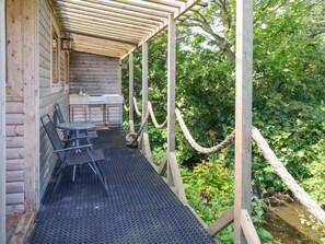 Una cabaña de madera con un porche cubierto, una mesa con sillas y un puente colgante sobre un arroyo.