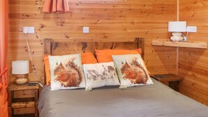A wooden cabin-style room with a bed, pillows, a nightstand, a lamp, and a window. - The Cabin (Amlwch)
