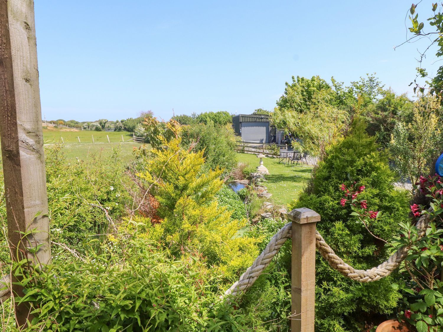 Un jardin avec une clôture en bois, un portail fait d’une corde et un bâtiment en arrière-plan.