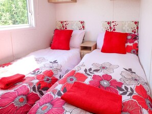Ein Schlafzimmer mit zwei Betten, jeweils mit einem blumigen Überwurf und roten Kissen, einem Nachttisch und einem Fenster mit Blick auf Grünflächen.