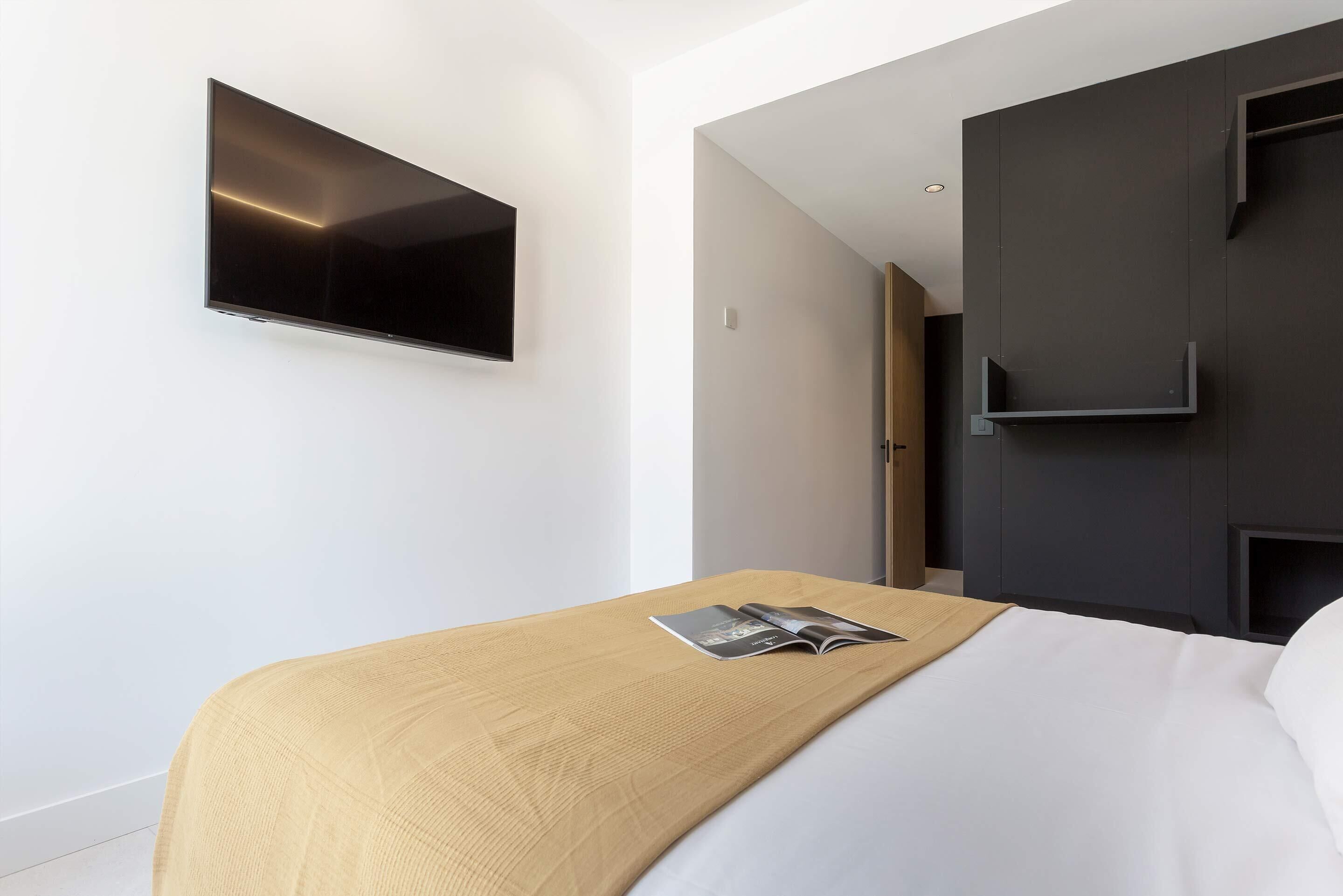 A modern hotel room with a flat-screen TV mounted on the wall, a bed with a wooden headboard, and a dark-colored wardrobe.