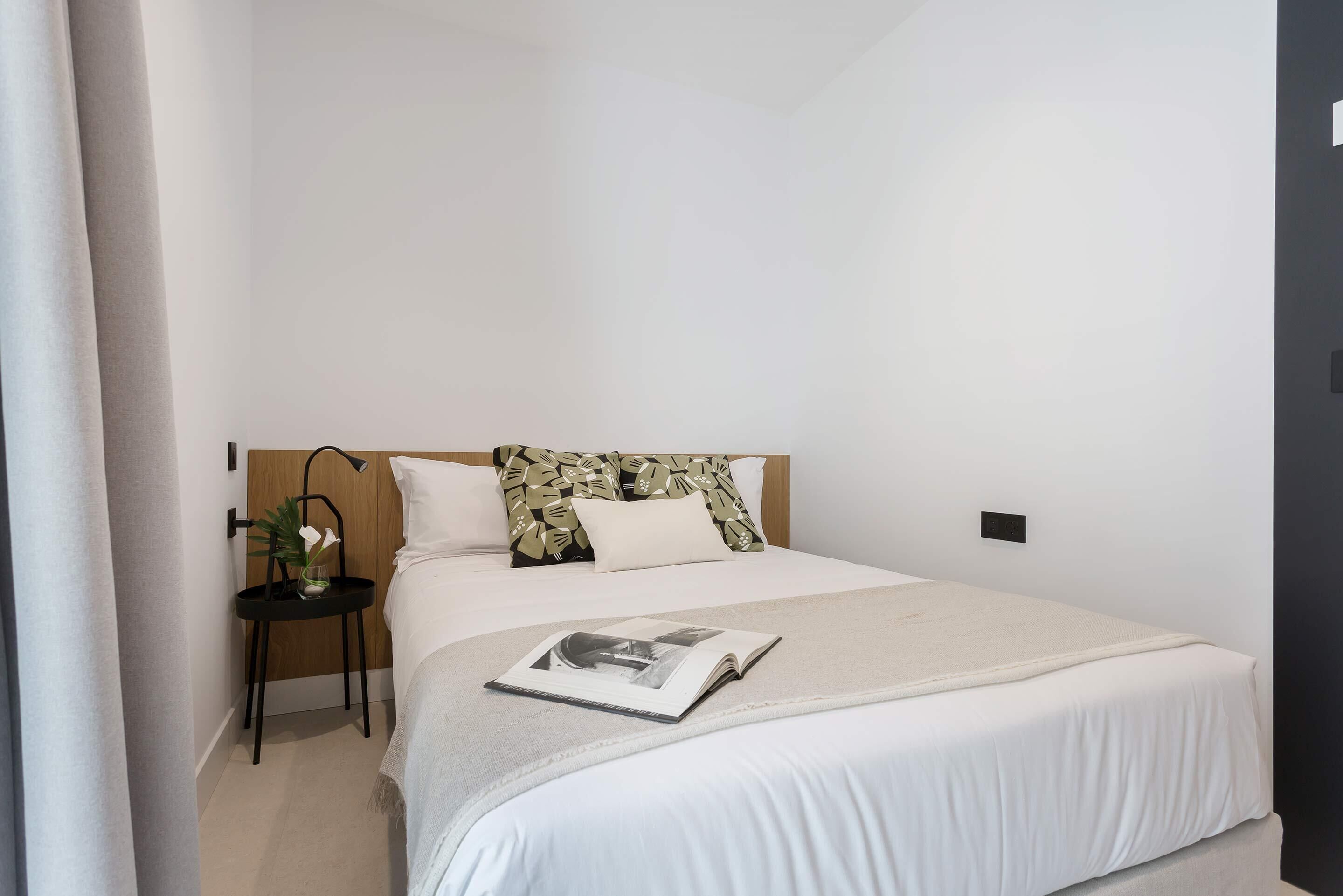 A neatly made bed with a white comforter and a book on top. There is a small side table with a plant and a lamp. The room has a simple, clean design with white walls and a wooden headboard.