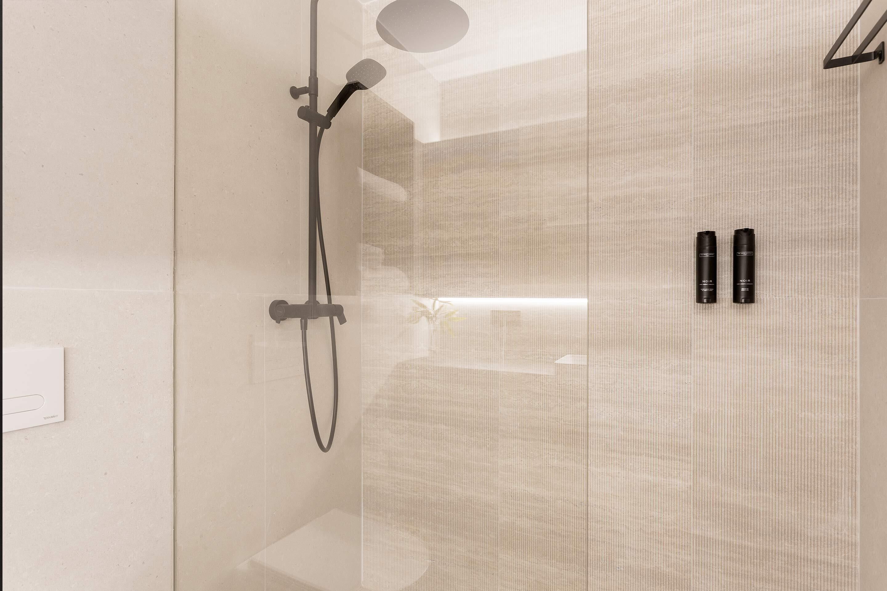 A modern shower with a handheld showerhead and a wall-mounted faucet, a glass shower enclosure, and two black bottles on the wall.