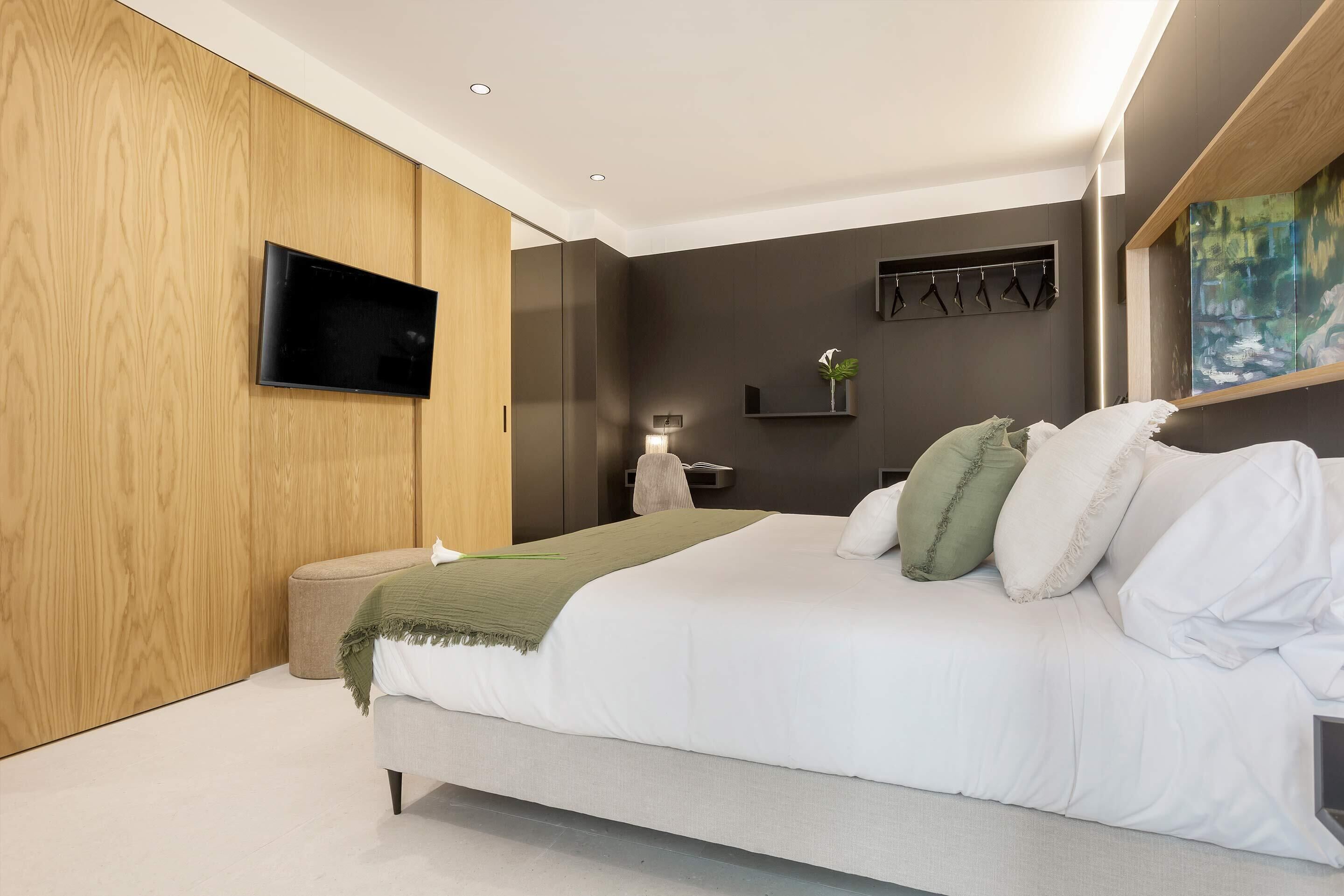 Un dormitorio moderno con una cama grande, un televisor de pantalla plana montado en la pared, una mesita de noche con una lámpara y un pequeño estante con una planta.