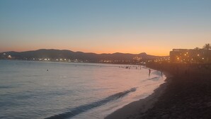 Una playa con un mar tranquilo, una luna creciente y luces de la ciudad a lo lejos.