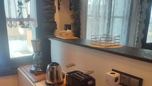 Una cocina con un tostador, hervidor y cafetera sobre la encimera, un estante pequeño con objetos decorativos y una ventana con cortinas.