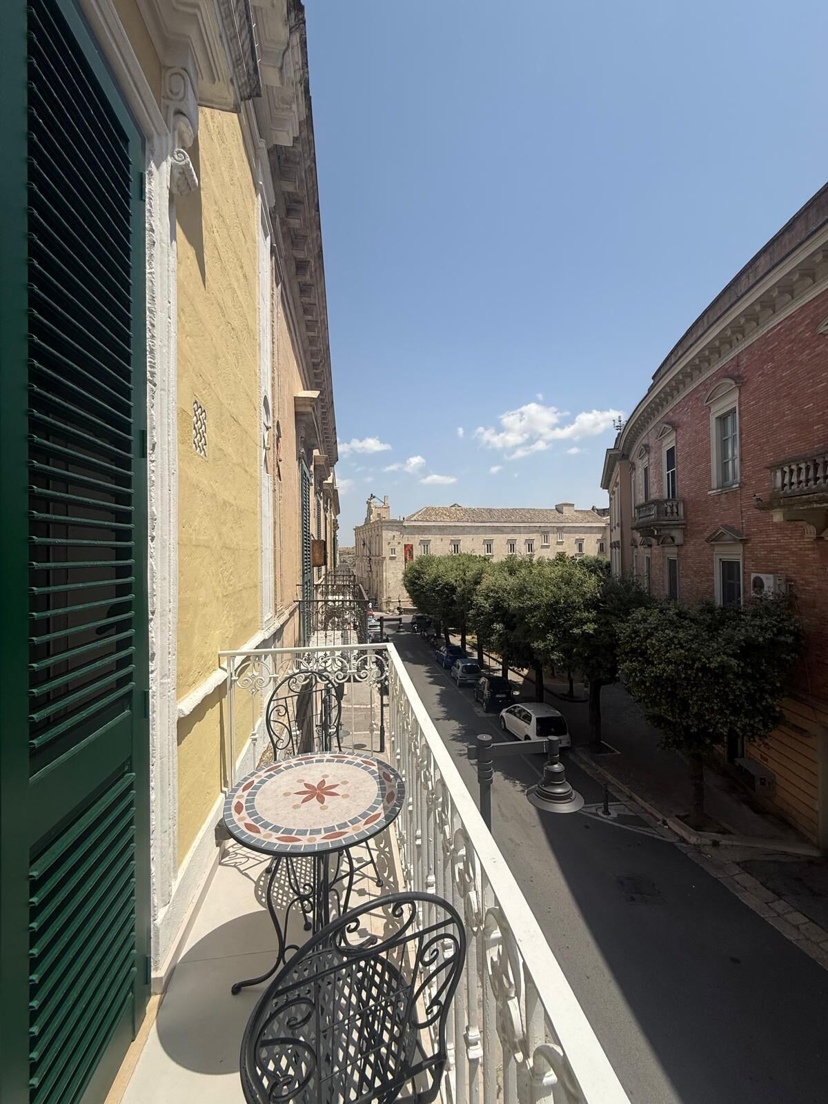 Un balcone con un tavolo e sedie piastrellati, vista su una strada con auto parcheggiate e edifici.