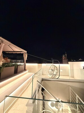 A rooftop terrace with a glass balustrade, a hanging chair, and a staircase leading to an upper level.