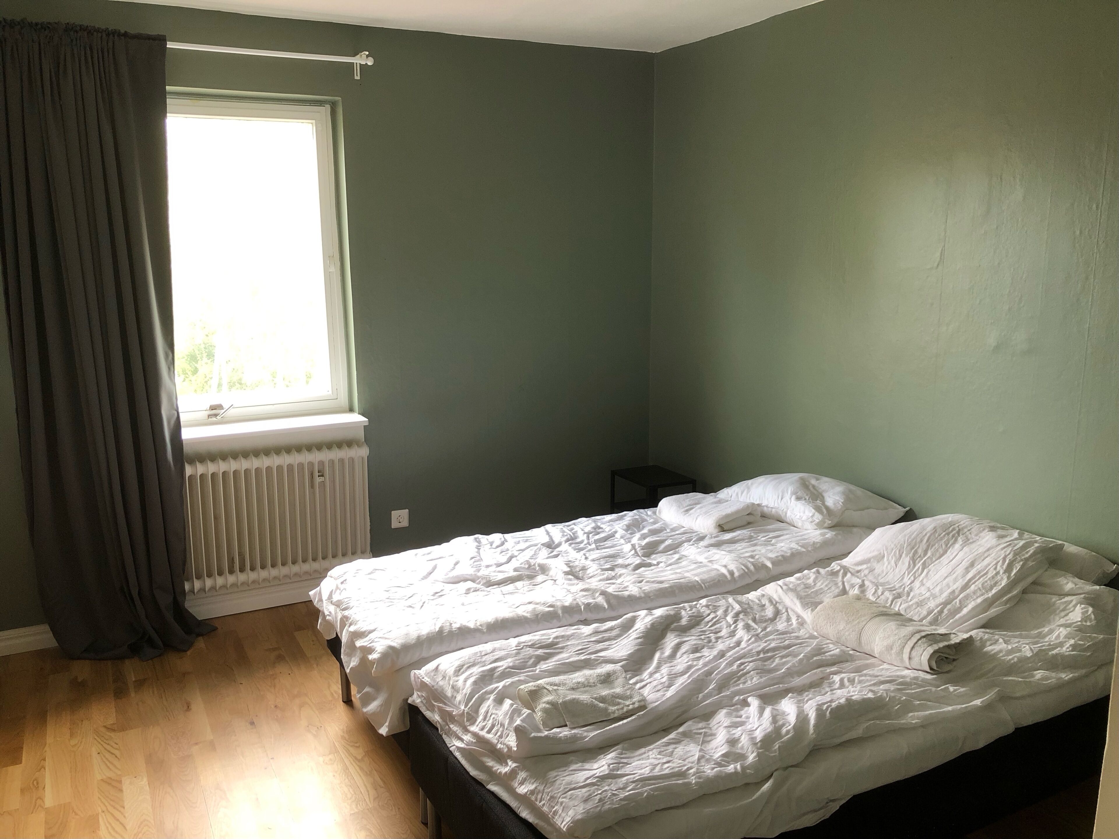 Una habitación con dos camas, una ventana con cortinas, un radiador y una mesita pequeña.