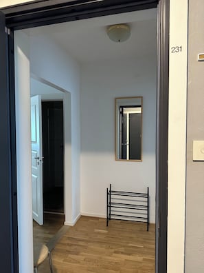 Un pasillo con una puerta que da a una habitación oscura, un espejo en la pared y un perchero metálico.