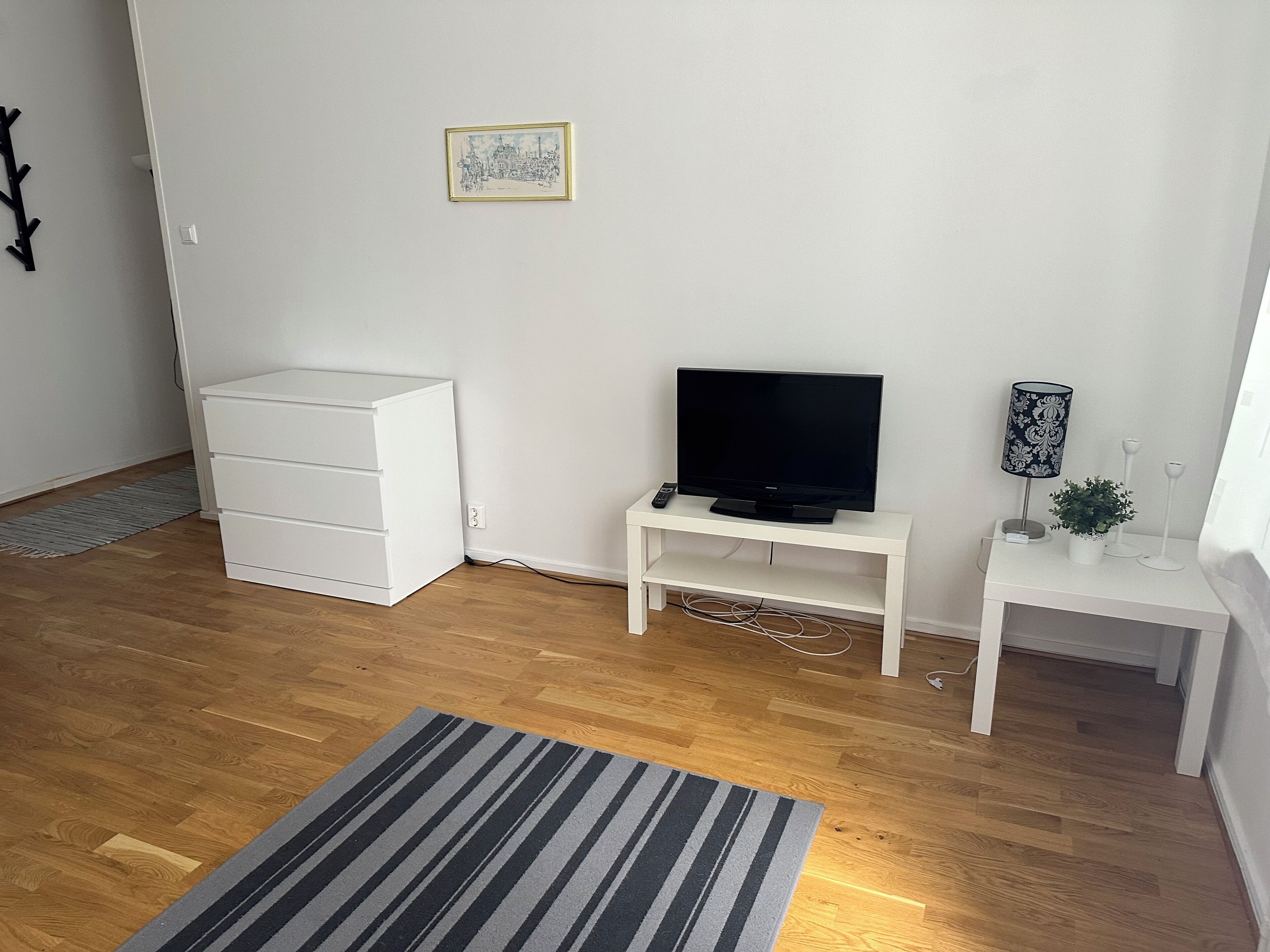 Une pièce avec un meuble blanc pour la télévision, un téléviseur à écran plat, une petite table blanche avec une lampe, un tableau encadré accroché au mur et un tapis à rayures sur le sol.