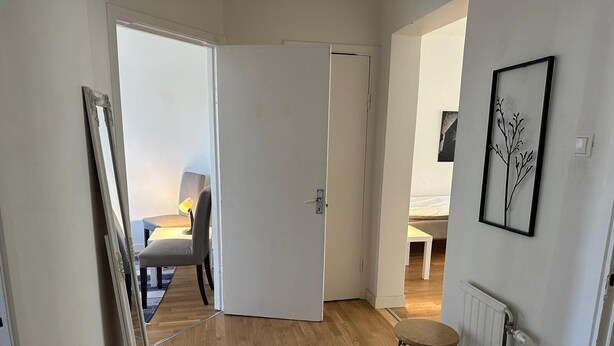 A hallway with a wooden floor, a patterned rug, a chair, a table with a lamp, and a bed in the background. - Mälartorget - Stay Smart (Västmanlands län)
