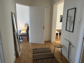 Ein Flur mit Holzboden, einem gemusterten Teppich, einem Stuhl, einem Tisch mit Lampe und einem Bett im Hintergrund.