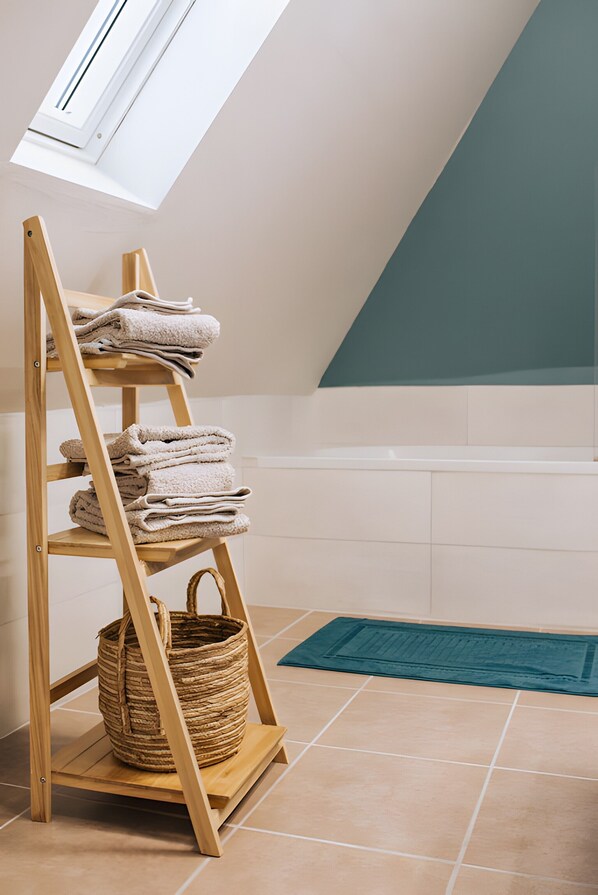 A wooden shelf with folded towels and a woven basket in a room with a sloped ceiling and a bathtub.