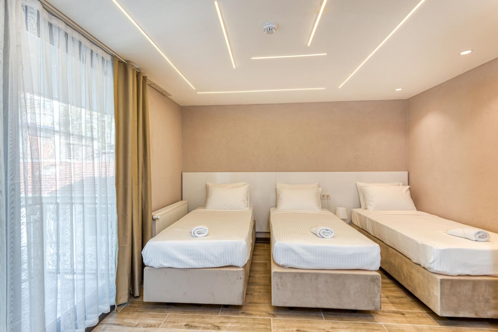 Habitación de hotel con dos camas individuales, un ventanal grande con cortinas translúcidas y un diseño moderno de techo.