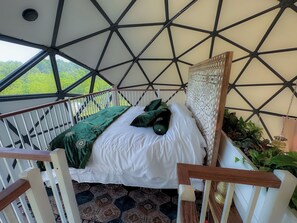 Una habitación con techo geométrico, cama con ropa de cama blanca y almohadas verdes, vista a zonas verdes exteriores y baranda con plantas.
