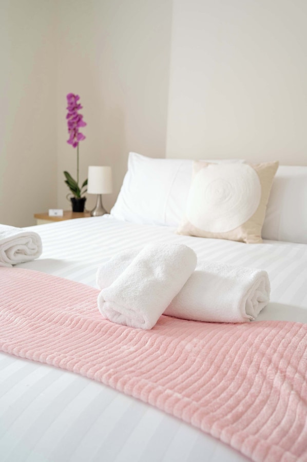 Una cama bien hecha con ropa de cama blanca y rosa, una toalla blanca enrollada, un plaid rosa y una maceta con flores moradas.