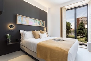 A modern bedroom with a large bed, a bedside table with a vase, a wall-mounted light, and a large window with a view of buildings and trees. - Elegant 3BD Apartment in Valladolid Solanilla II (Valladolid)