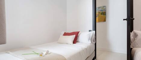 Una cama individual con ropa de cama blanca y una almohada roja, un adorno verde, y un cuadro en la pared.