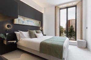 A modern bedroom with a large bed, a side table with a plant, a painting above the bed, and a window with a view of buildings. - Bright Apartment in the Heart of Valladolid Solanilla IV (Valladolid)