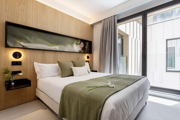 A modern bedroom with a large bed, a wall-mounted painting, bedside tables, and a view of the outside.