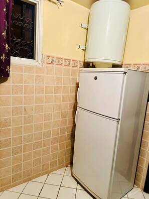 Een compacte keuken met een koelkast, een waterdispenser en een raam met decoratieve metalen tralies.