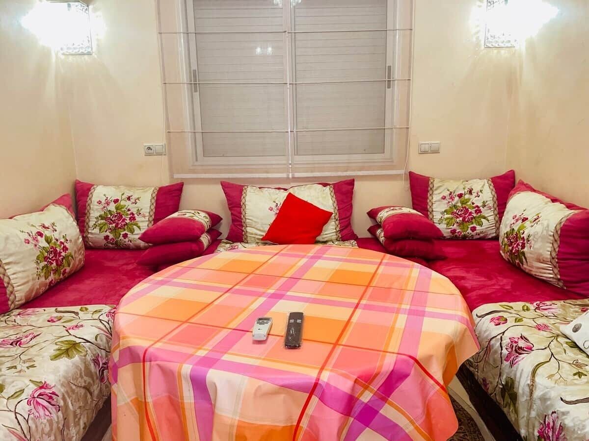 A room with a central bed, two side beds, a window with blinds, and a table with two remotes.