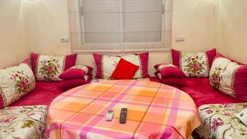 A room with a central bed, two side beds, a window with blinds, and a table with two remotes.