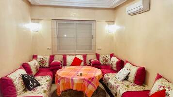 A room with a sofa set, a round table, a window, and an air conditioning unit.