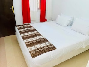 Ein Bett mit weißer Bettwäsche und einer gemusterten Decke, rote Vorhänge und eine Tür mit gelbem Griff.