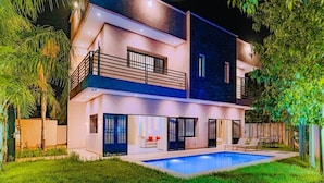 Une maison moderne à deux étages, dotée d’une piscine, de balcons et d’un éclairage extérieur soigné.