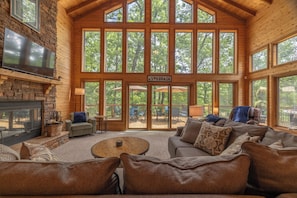 A spacious living area with a stone fireplace, a flat-screen TV, and large windows offering views of the outdoors. - Sunset Haven by Innsbrook Vacations! (Innsbrook)