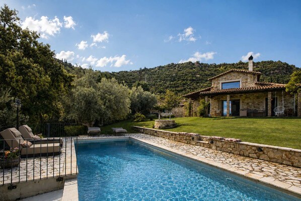 Une piscine avec une terrasse en pierre, une maison en pierre avec un toit en tuiles et une zone herbeuse avec des arbres.