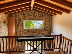 A stone-walled room with a piano, a window with a view of trees and mountains, and a wooden ceiling.