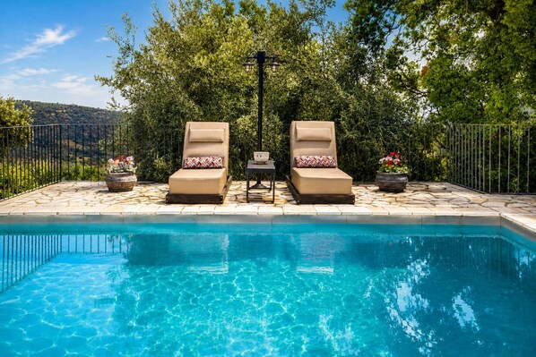 A swimming pool with two lounge chairs and a table, surrounded by a fence and trees.