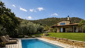 Une piscine aux bords recouverts de pierres, une maison à la façade en pierre et une pelouse verdoyante.
