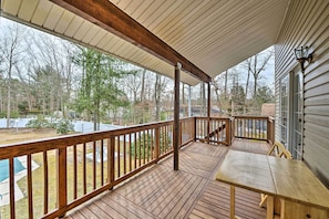 A covered wooden deck with a table and chairs, overlooking a backyard with trees and a swimming pool.