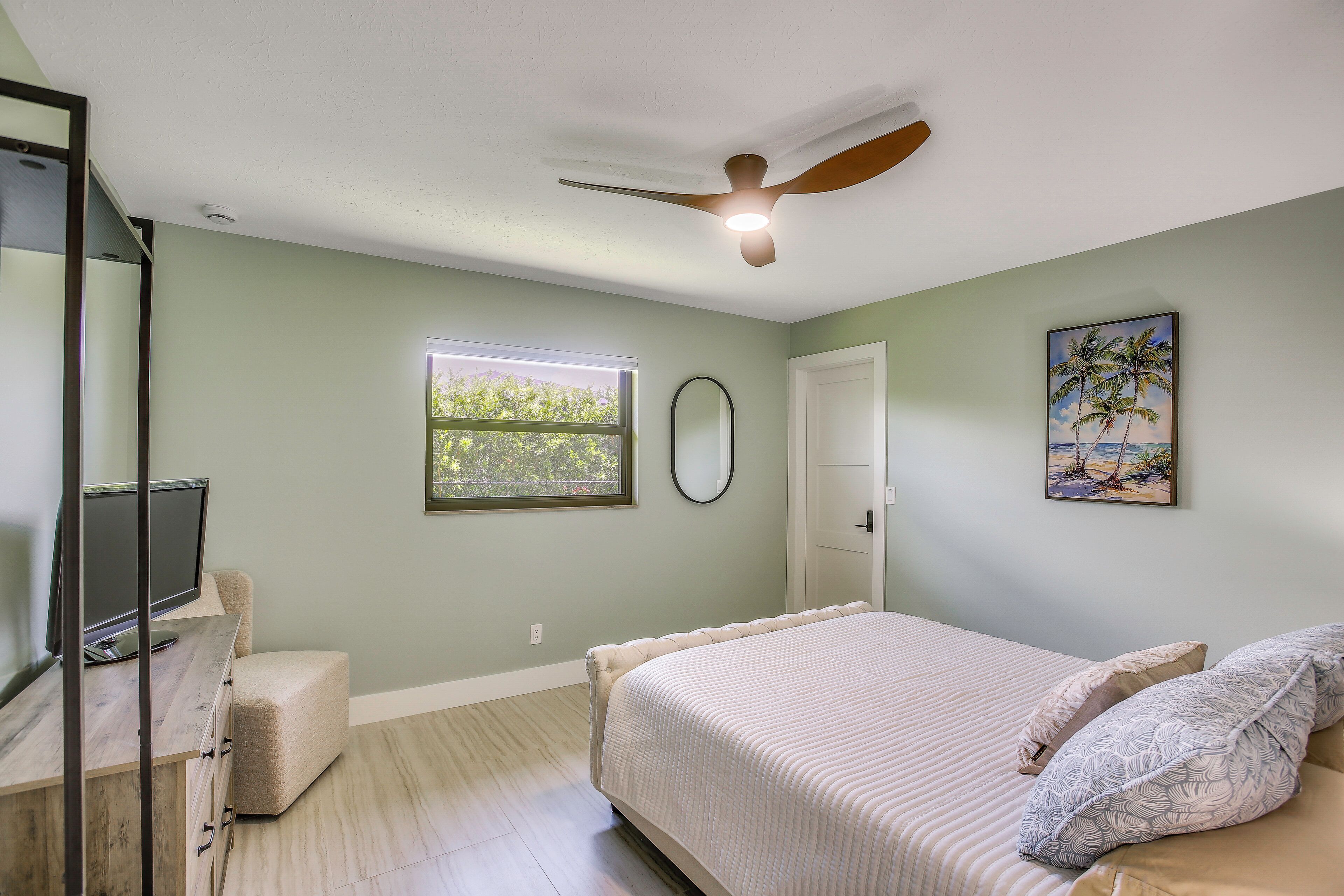 A bedroom with a bed, a desk with a computer, a ceiling fan, a window, and a door.