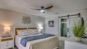 Ein Schlafzimmer mit Bett, Nachttischen, Deckenventilator, einer Topfpflanze und einer Schiebetür zum Badezimmer.