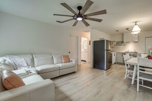 A modern living room with a white sofa, a dining table with chairs, a kitchen area with a refrigerator, and a ceiling fan.