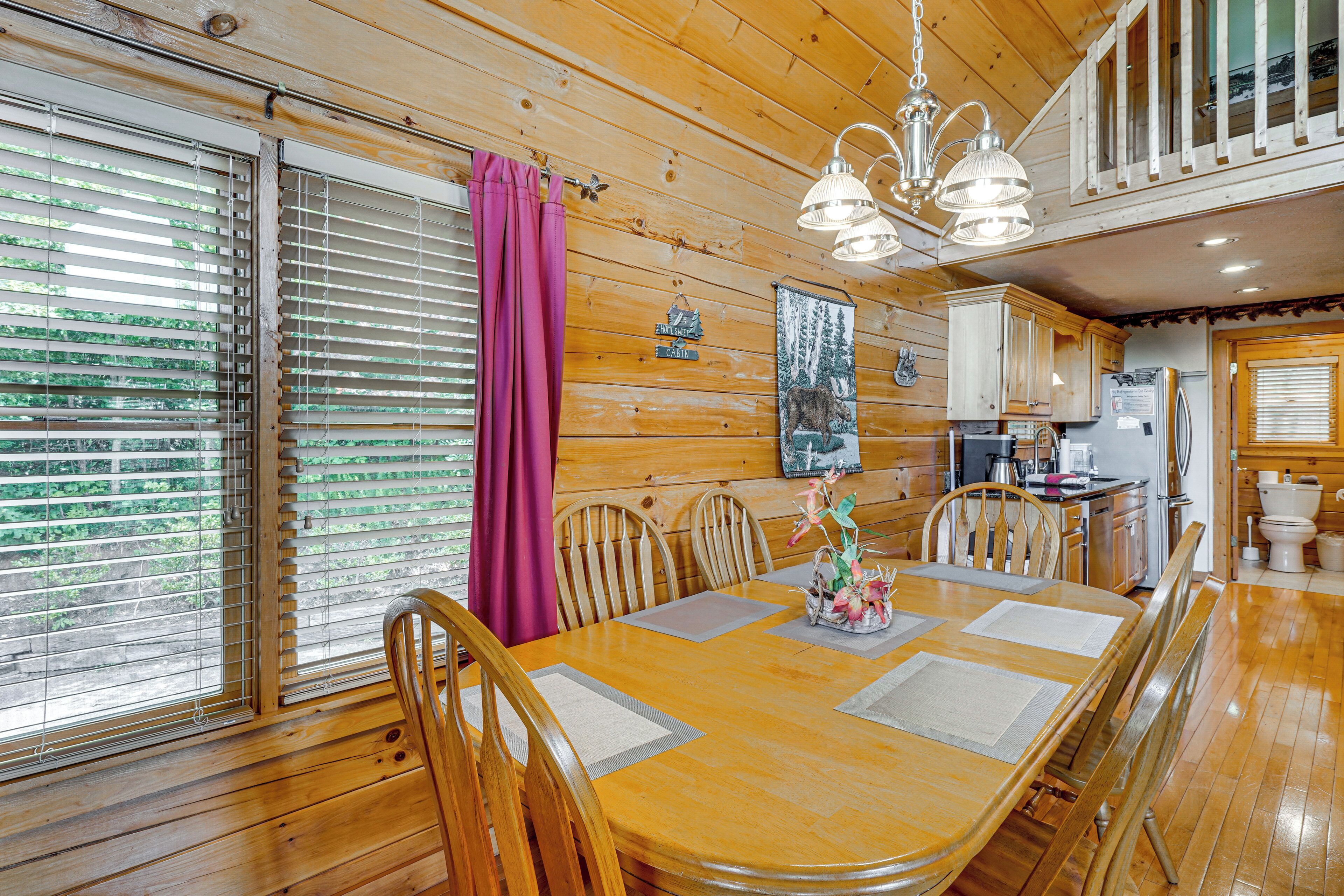 Interior de una cabaña de madera con una mesa de comedor, sillas, una zona de cocina y un ventanal con persianas.