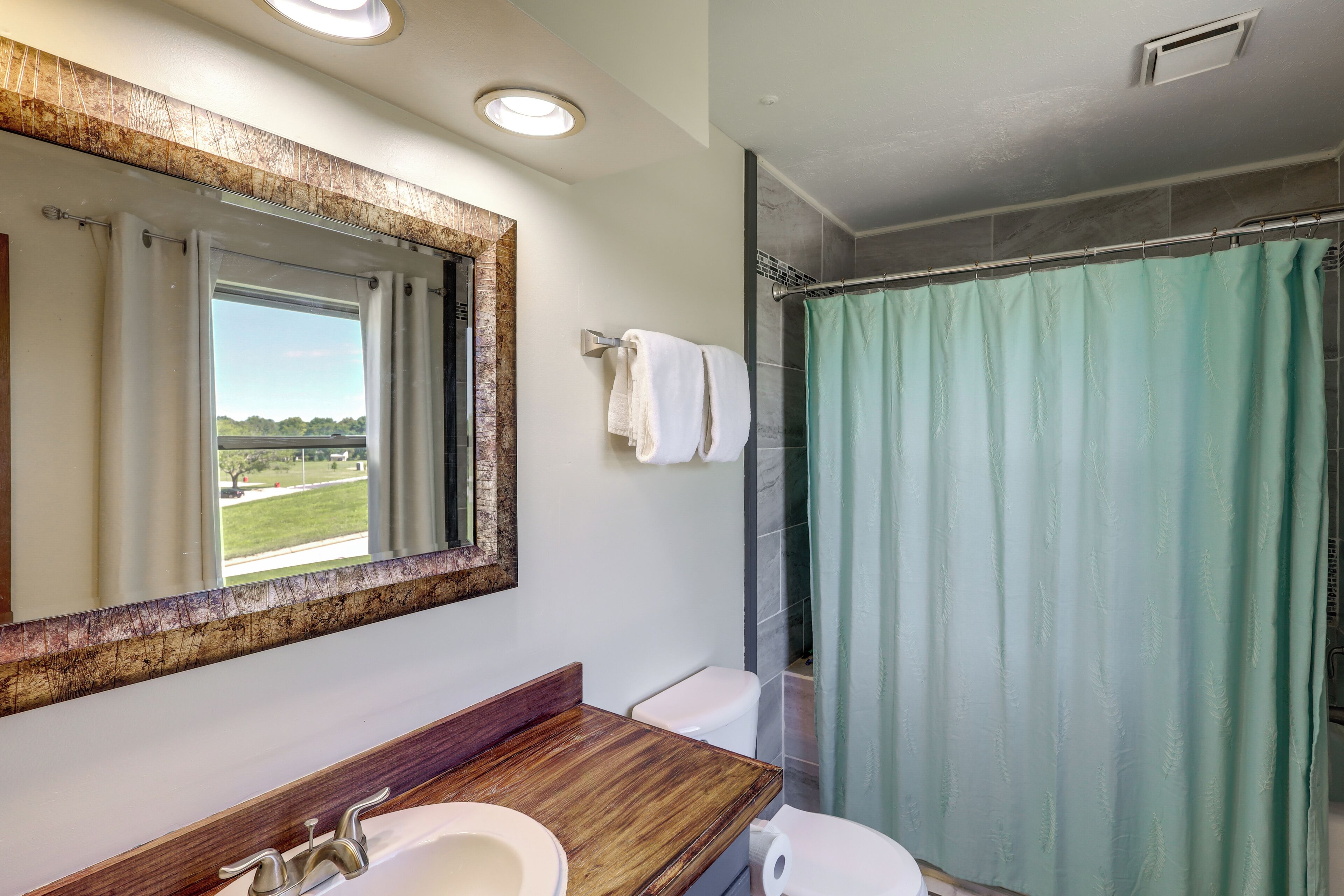 Ein Badezimmer mit einem großen Spiegel, einem hölzernen Waschtisch, einer Toilette, einer Duschvorhang und Blick ins Freie.