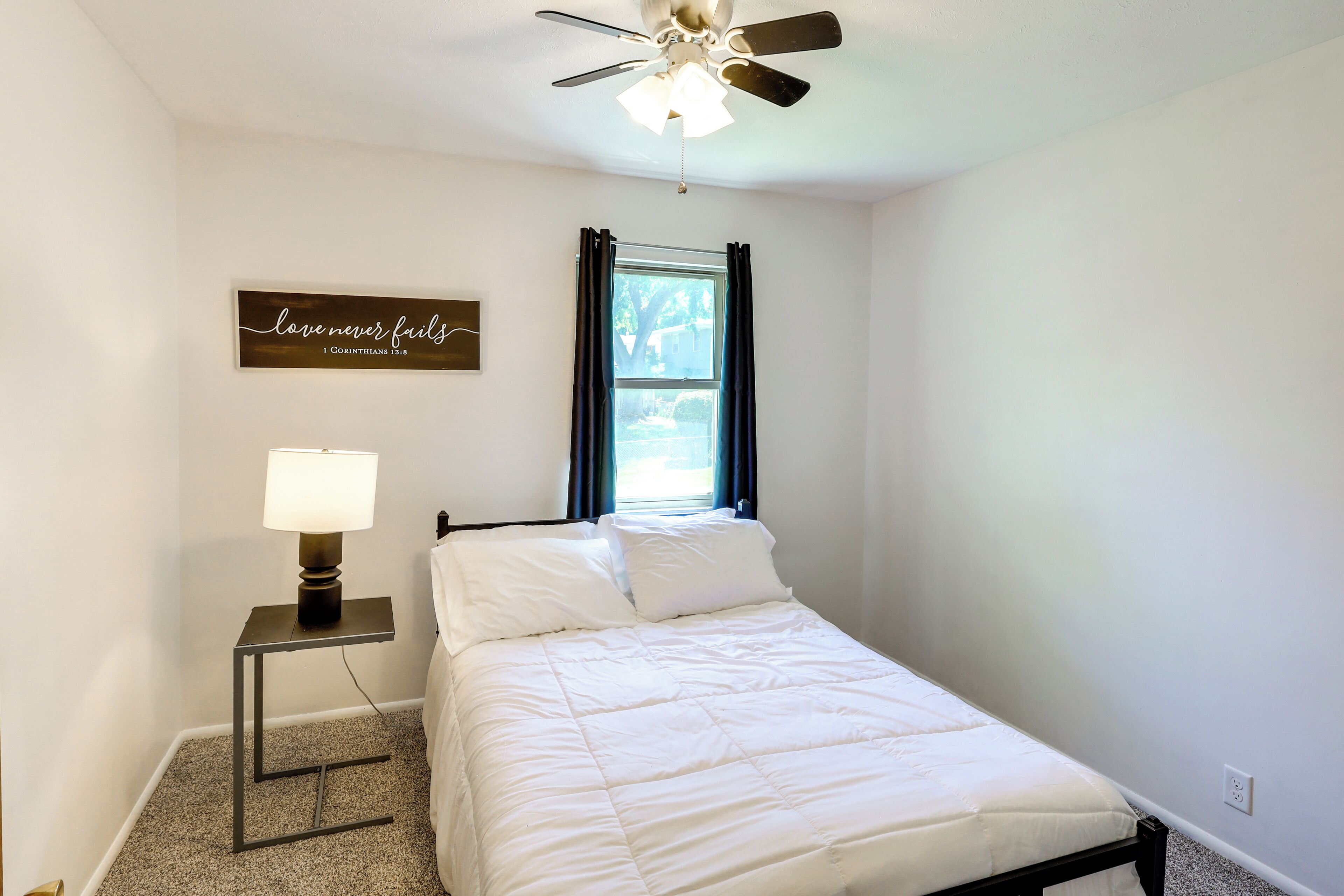 A bedroom with a bed, a nightstand with a lamp, a ceiling fan, a window with curtains, and a motivational quote on the wall.