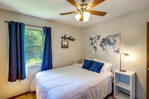 Un dormitorio con cama, ventilador de techo, ventana con cortinas, pared con un cuadro y la palabra 'adventure', y mesita de noche con lámpara.