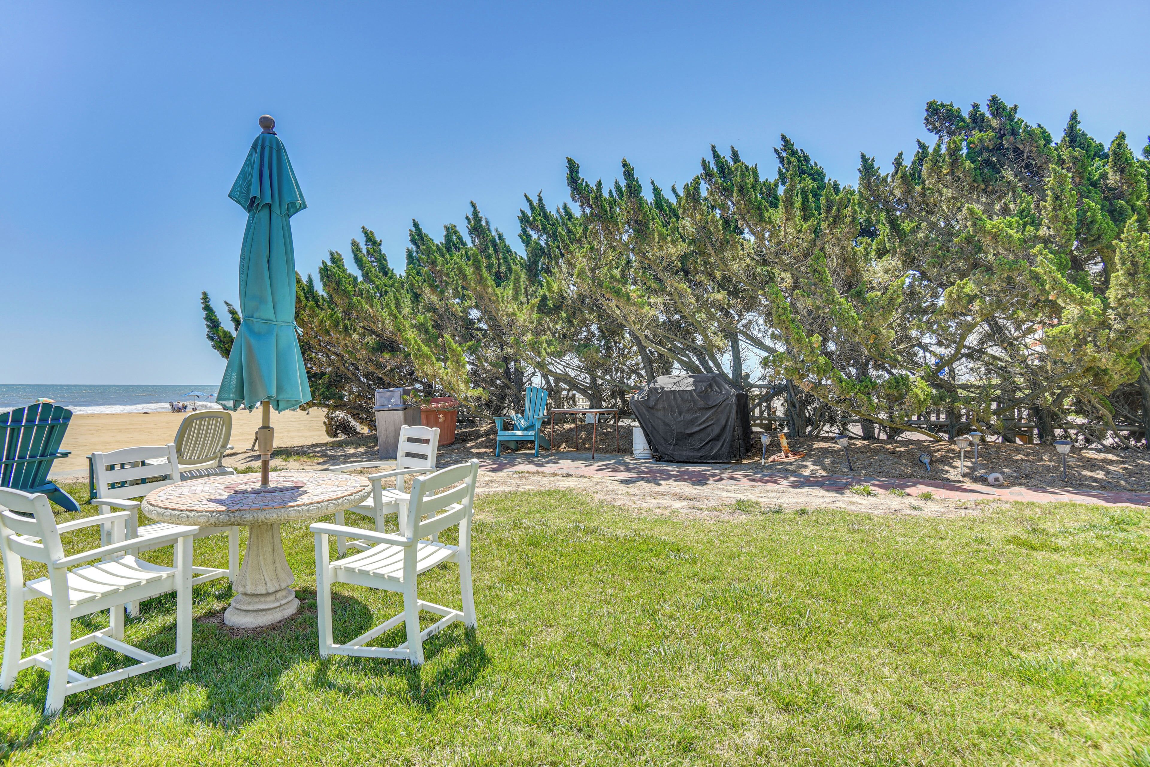A patio with outdoor furniture, a table, and chairs, a blue umbrella, and a view of the beach and trees.