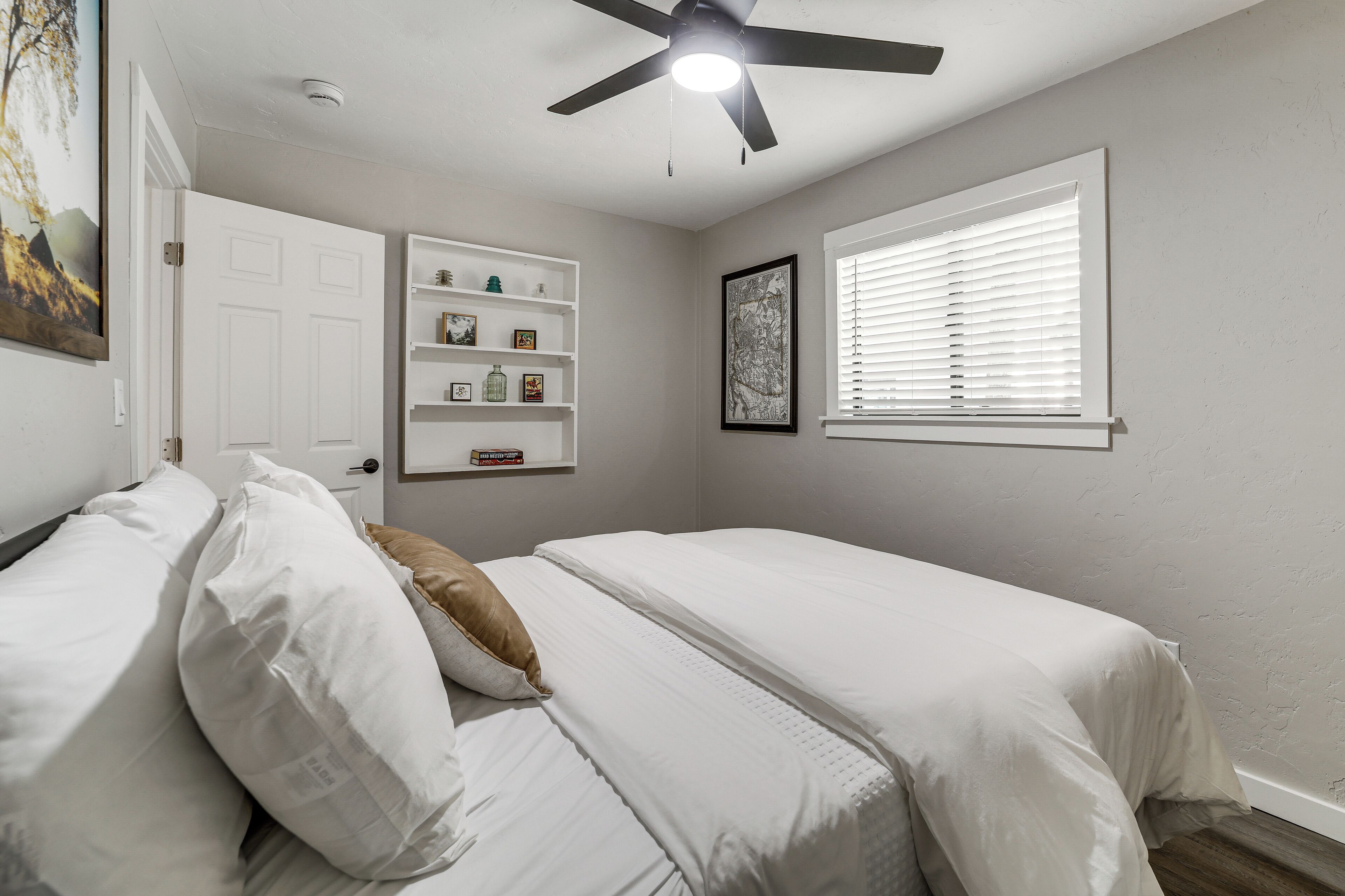 A bedroom with a bed, a ceiling fan, a window with blinds, a door, and a shelf with decorative items.